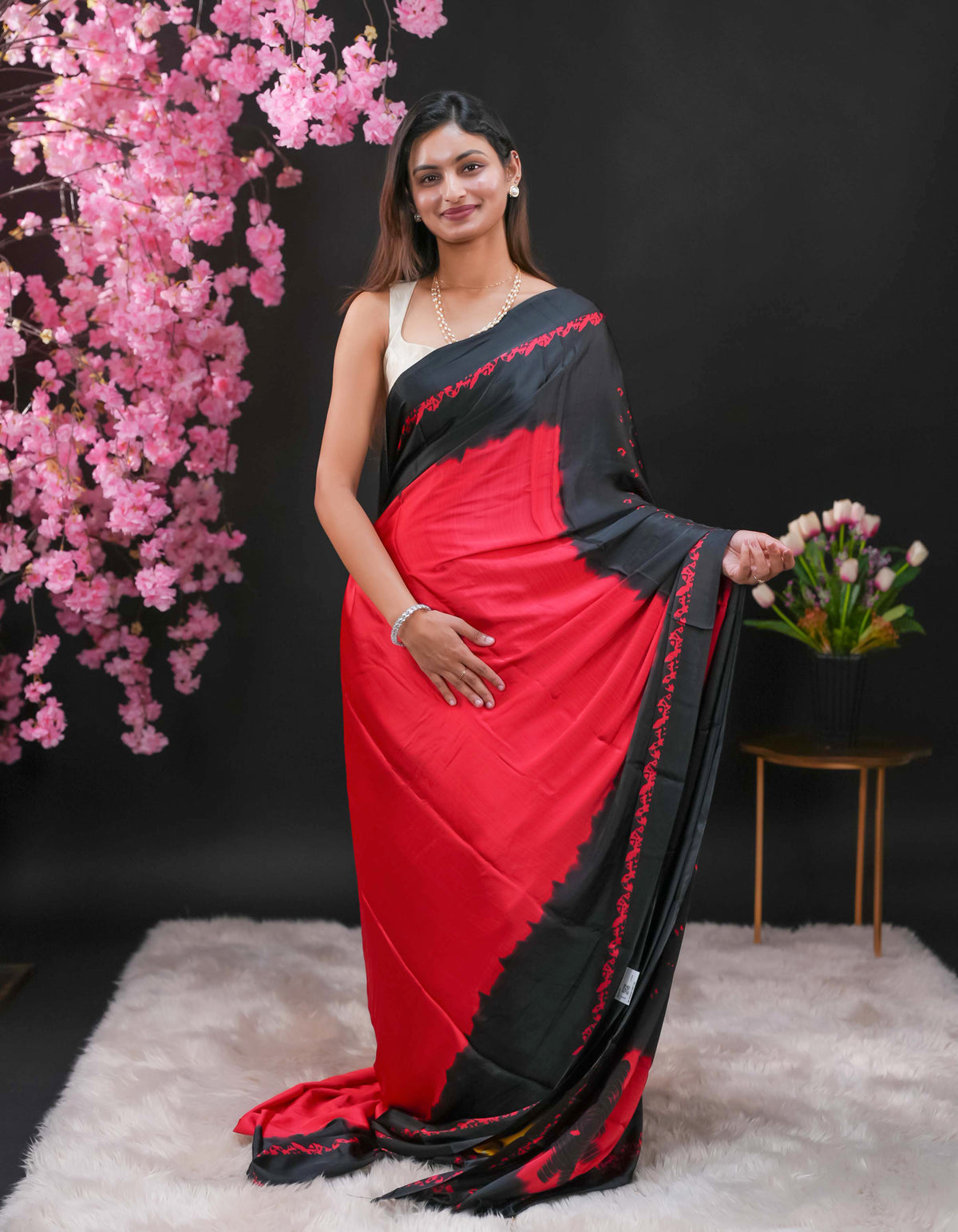 Black coloured modal satin saree featuring a plain Red coloured body, complemented by a double shaded border and a Bandhani designed pallu with jhumka tassels. Paired with a matching contrast coloured blouse piece.