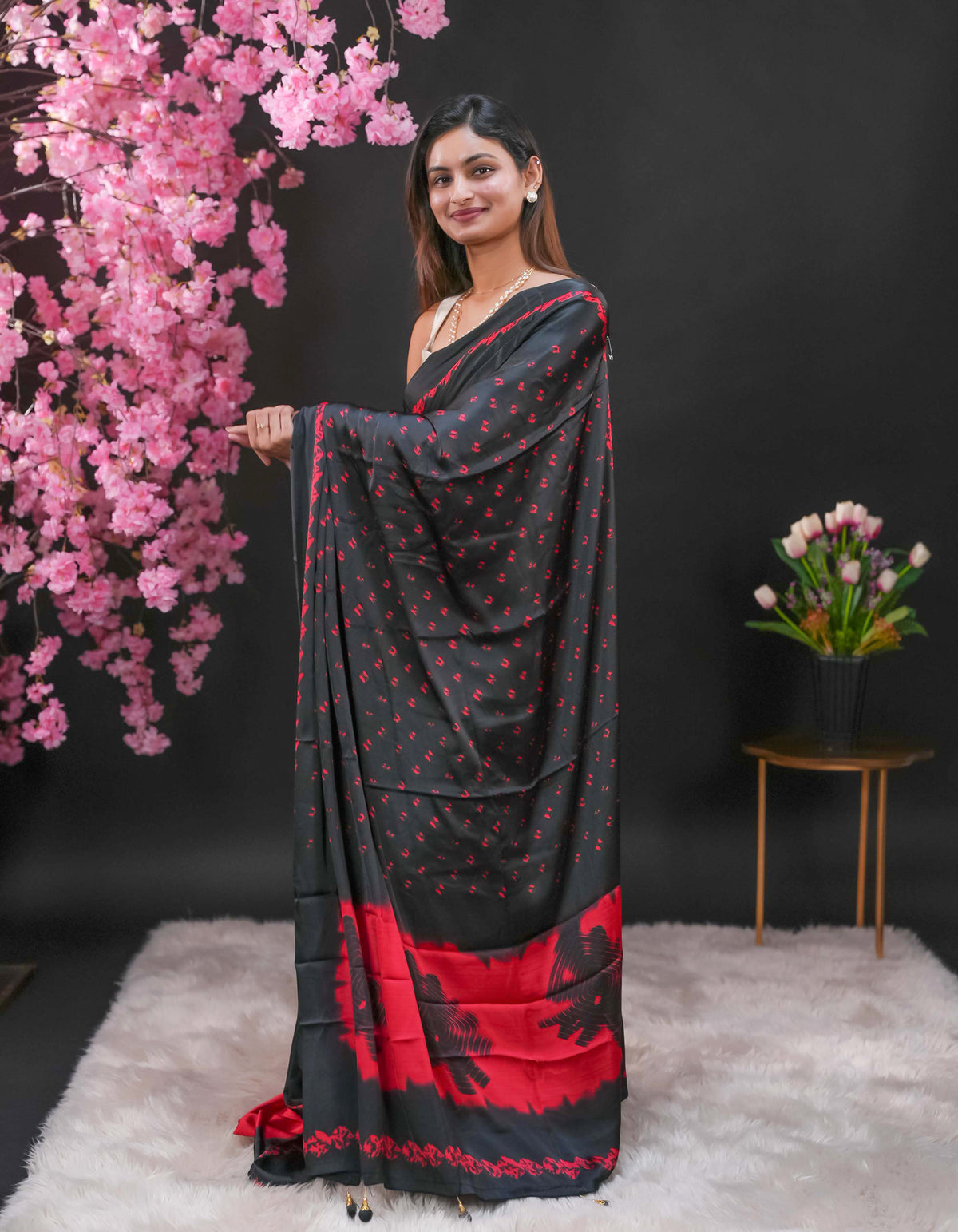Black coloured modal satin saree featuring a plain Red coloured body, complemented by a double shaded border and a Bandhani designed pallu with jhumka tassels. Paired with a matching contrast coloured blouse piece.