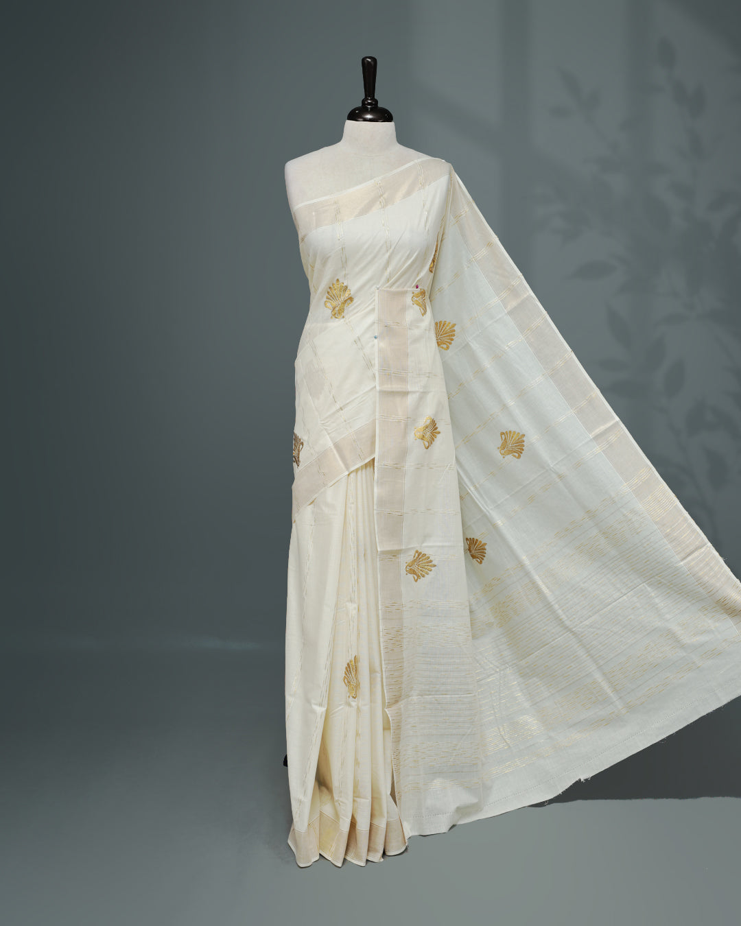 Cream colour Cotton kerala saree featuring Gold Embroidery strips with motifs on the body, complemented by a gold zari Border, paired with a matching blouse piece.