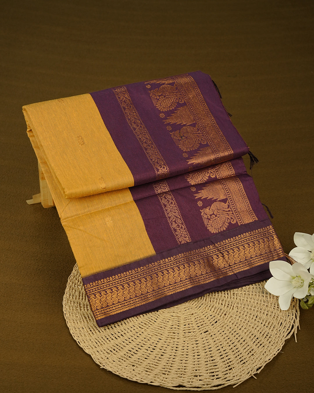 Mustard Yellow Colour Kalyani cotton saree featuring delicate antique zari floral butta motifs on the body, complemented by a Purple contrast pallu with a rich antique zari peacock border, paired with a matching blouse piece.
