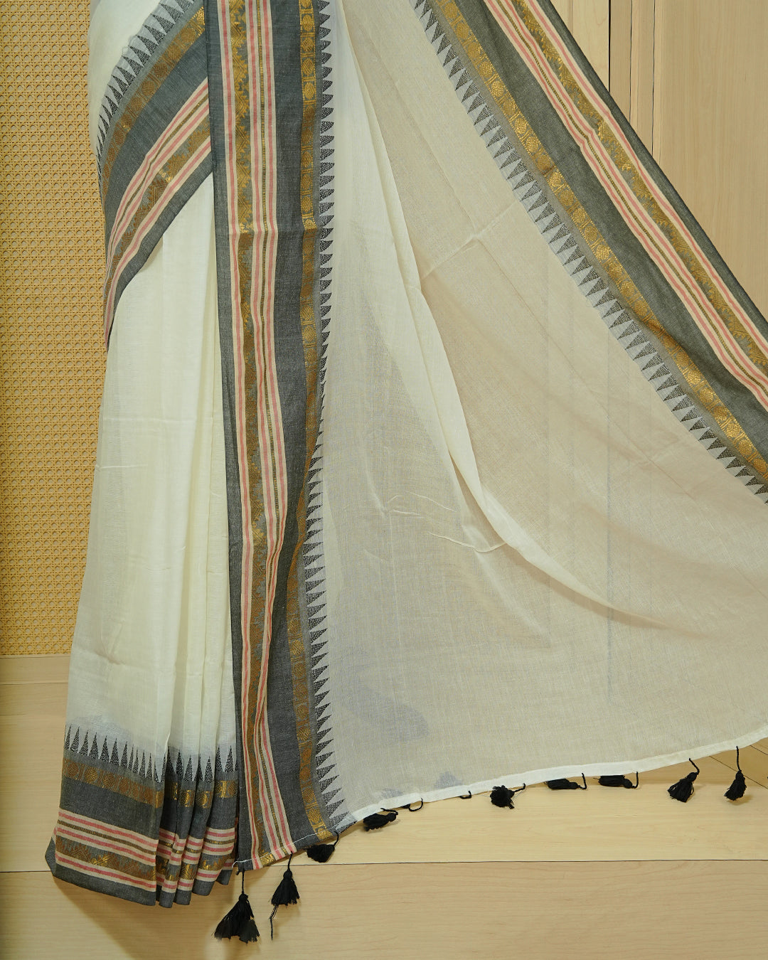 Cream Mul Cotton Saree featuring an elegant plain body, complemented by a grey contrast border with gold zari and subtle Red stripe detailing, finished with black tassels.