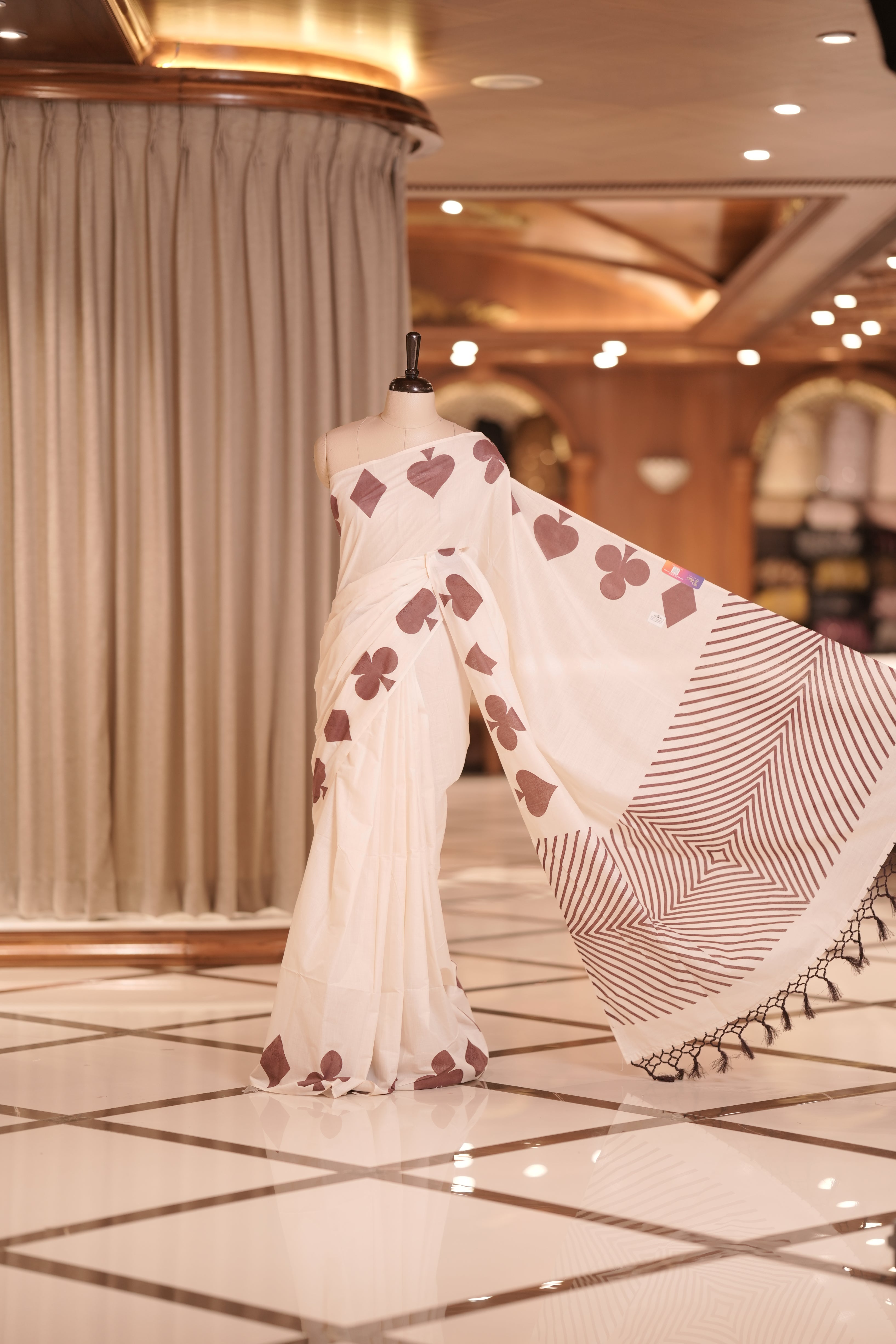 Off white colour kerala cotton saree adorned with brown colour card suit motifs , The pallu features striking zigzag patterns with tassel detailing, creating a perfect fusion of tradition and modern sophistication.