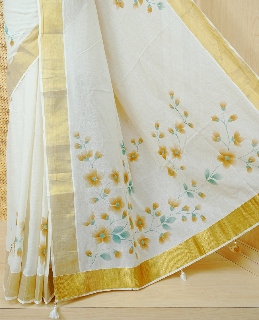 Cream Colour Cotton Kerala Saree Featuring Digital Floral Prints Across The Body, Enhanced With A Gold Zari Border, Traditional Gold Zari Pallu, Finished With Light Brown & White Tassels And A Matching Blouse Piece.