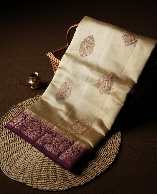 Cream Colour Banaras Soft Silk Saree with Gold Zari Oval Leaf Buttas, Intricate Multi-Layered Floral Border, Grand Pallu with Intricate Brocade Motifs in Deep Maroon and Gold Tones, and Matching Blouse.