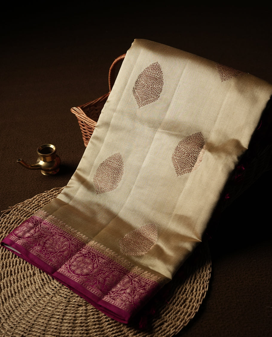 Cream Colour Banaras Soft Silk Saree with Gold and Rose Gold Zari Textured Diamond-Shaped Buttas, Simple Decorative Pallu Border, Grand Pallu with Ornate Floral and Circular Motifs in Magenta and Gold Tones, and Matching Bright Magenta Blouse.
