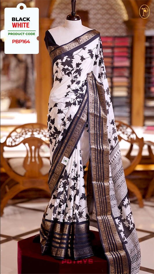 White colour semi tussar silk saree adorned with black floral prints, highlighted by a traditional border. Paired with a plain black blouse featuring matching border and a beautifully printed pallu for classic elegance.
