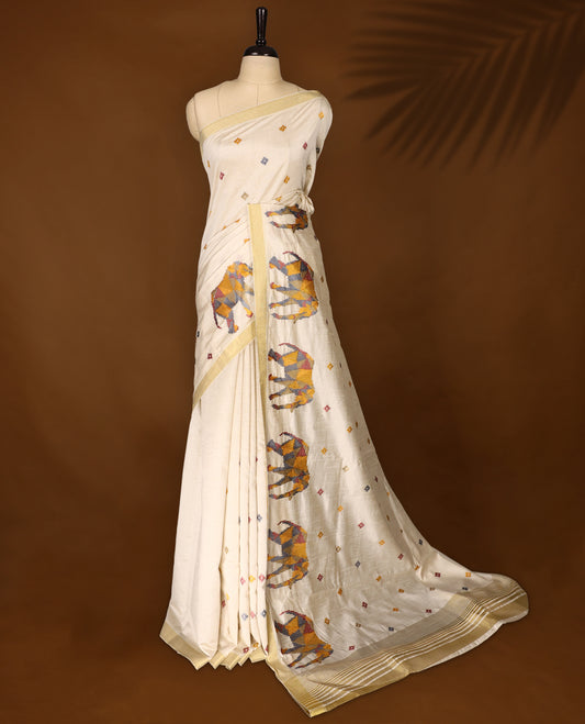 Cream colour Tussar saree adorned with multicolour geometric elephant motifs and delicate thread buttas across the body, enhanced with a subtle gold zari border, paired with a matching blouse.