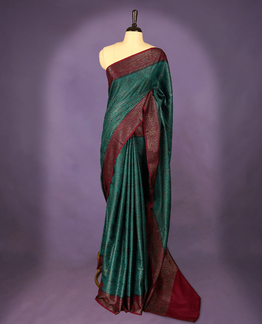 Dark aqua green colour semi Banaras Katan saree featuring an antique zari jacquard body, complemented by a contrasting traditional border with a floral kodi design, paired with a matching blouse.