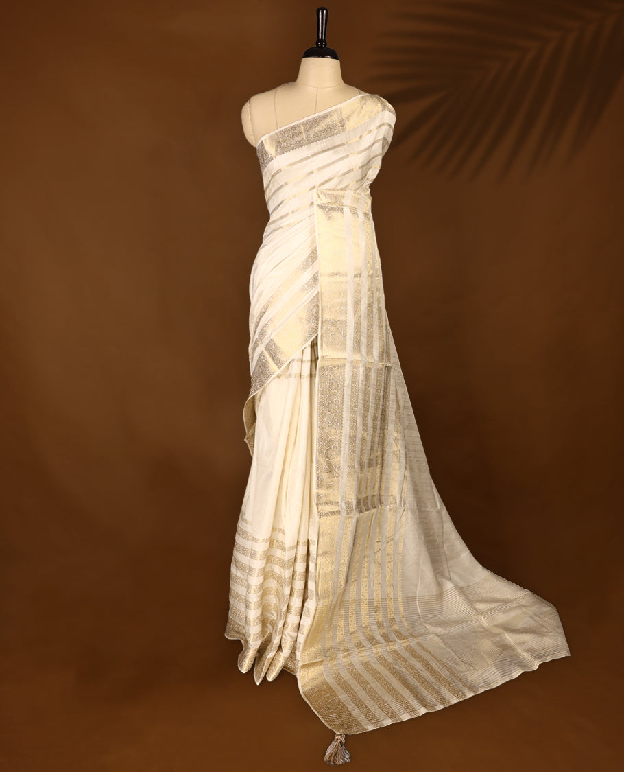 Ivory white Colour Tussar Kerala saree with a sheer, smooth body and soft golden sheen, Accented with fine zari stripes and an intricate floral zari border,Comes paired with a coordinated matching blouse piece.