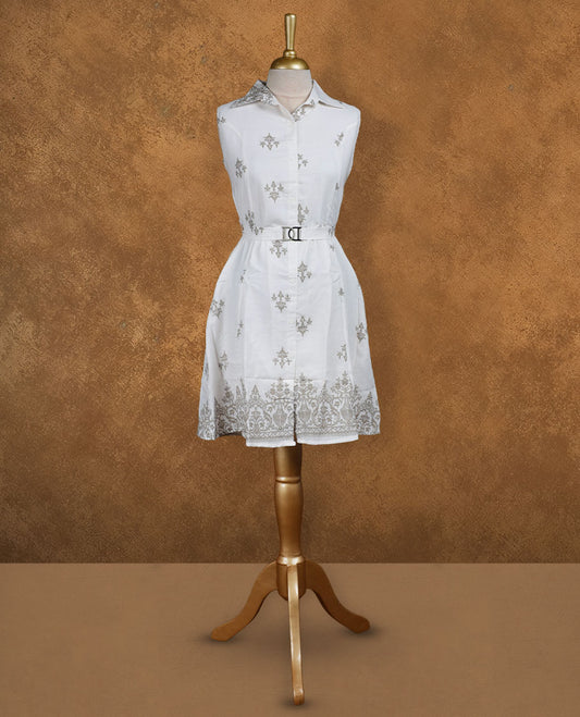 White sleeveless shirt dress with subtle grey ethnic motifs, front button placket, matching waist belt with metal buckle, and ornate border print at the hem.