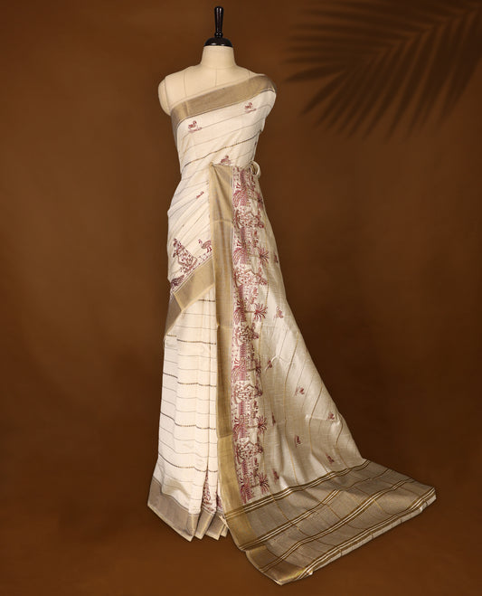 Cream colour Soft Tussar saree featuring fine golden woven stripes across the body, accentuated with maroon village and temple motifs on the pallu, finished with a rich golden zari border, paired with a matching blouse.