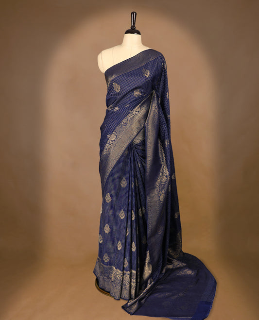 Navy Blue coloured semi Banaras georgette saree featuring golden Zari checks with floral motifs on the body, complemented by a self coloured traditional border with floral kodi design and a matching blouse.