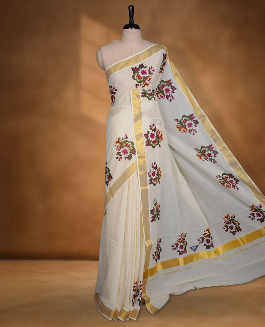 Off-white colour cotton Kerala saree with a classic gold kasavu border, adorned with multi colour floral motifs across the body and pallu, and paired with a matching blouse.