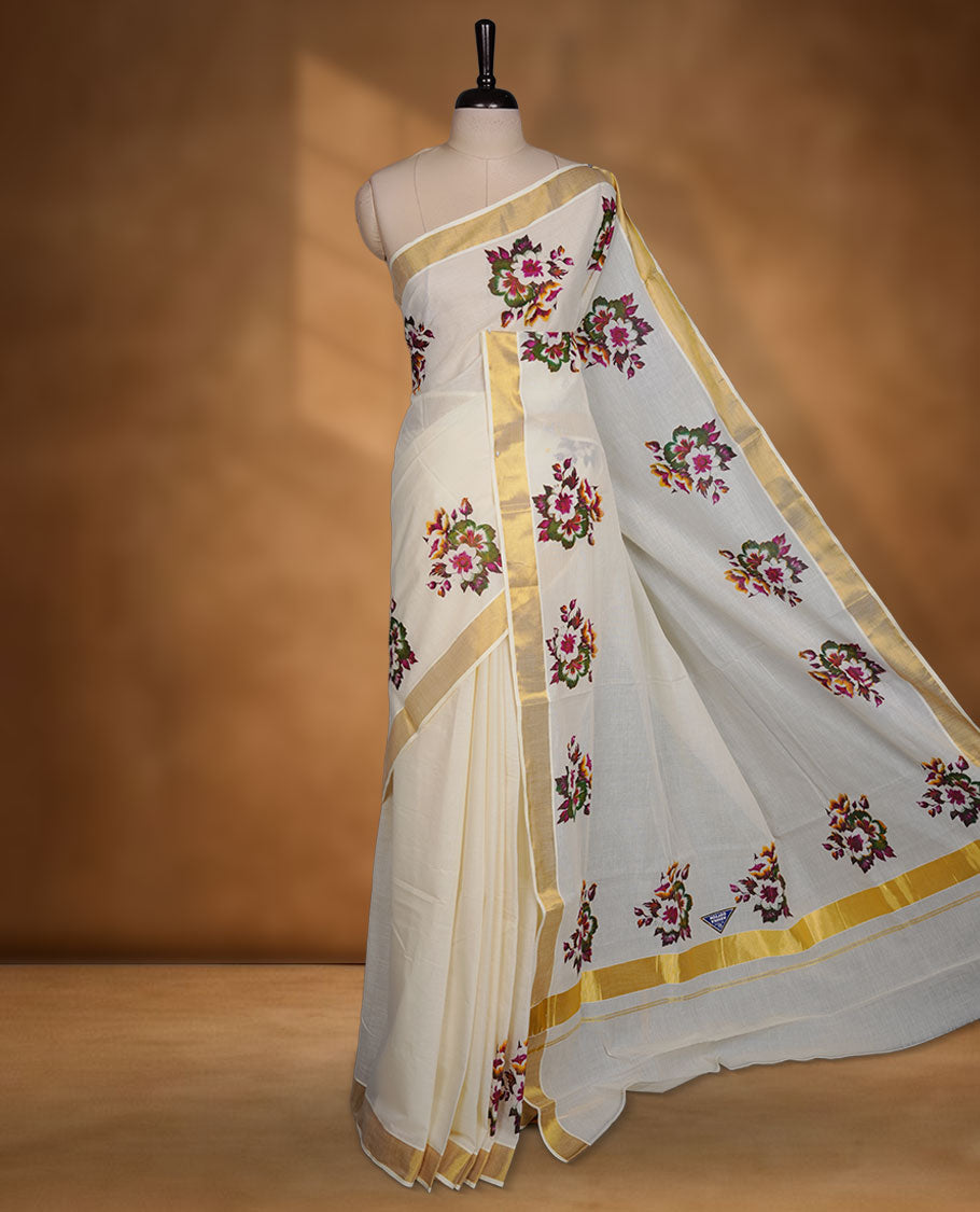 Off-white colour cotton Kerala saree with a classic gold kasavu border, adorned with multi colour floral motifs across the body and pallu, and paired with a matching blouse.