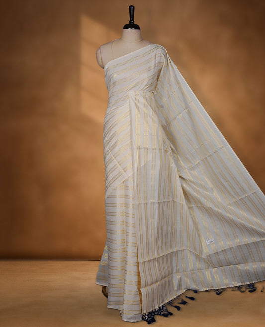 Off-white colour cotton Kerala saree featuring subtle gold zari stripe detailing across the body, finished with elegant tassels on the pallu, and paired with a matching blouse.