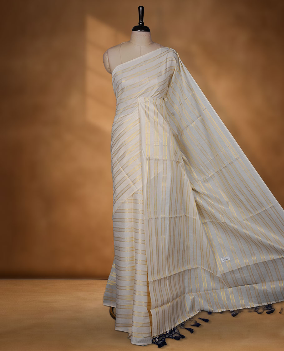 Off-white colour cotton Kerala saree featuring subtle gold zari stripe detailing across the body, finished with elegant tassels on the pallu, and paired with a matching blouse.