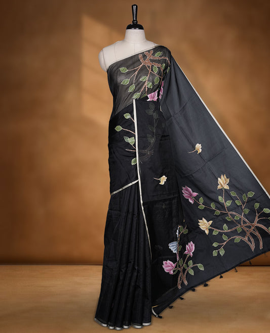 Black colour semi tusser silk saree featuring delicate floral embroidery motifs across the body with subtle contrast detailing on the border, paired with a matching embroidered blouse.