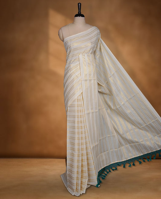 Off-white colour cotton Kerala saree featuring subtle gold zari stripe detailing across the body, finished with elegant green tassels on the pallu, and paired with a matching blouse.