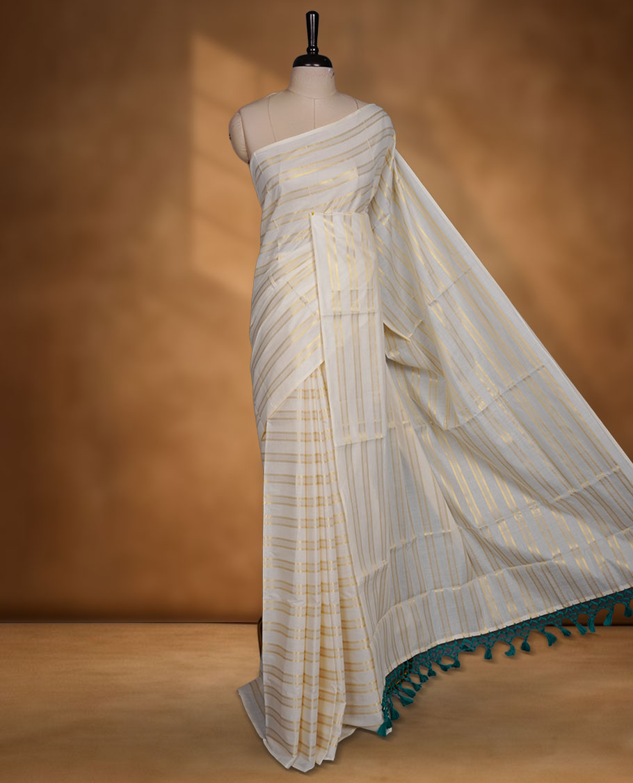 Off-white colour cotton Kerala saree featuring subtle gold zari stripe detailing across the body, finished with elegant green tassels on the pallu, and paired with a matching blouse.