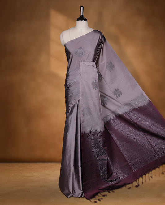 Reddish gray colour semi soft silk saree adorned with delicate woven floral motifs on the body, complemented by a contrast deep maroon colour woven pallu with intricate detailing and tassel accents without border & Comes with a matching blouse piece.