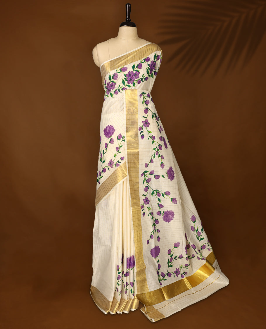 Elegant Off white colour cotton Kerala saree with hand-painted purple floral motifs, highlighted by a rich golden zari border and pallu paired with matching blouse.