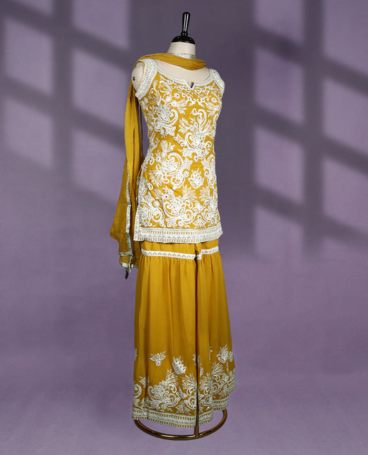 Mustard colour sharara set features a beautifully embellished sweetheart neckline with cutout detail and sleeveless design, adorned with intricate white embroidery throughout the kurti and hem of the flared sharara pants, paired with a matching dupatta