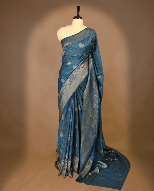 Blue coloured semi Banaras georgette saree featuring golden Zari checks with floral motifs on the body, complemented by a self coloured traditional border with floral kodi design and a matching blouse.