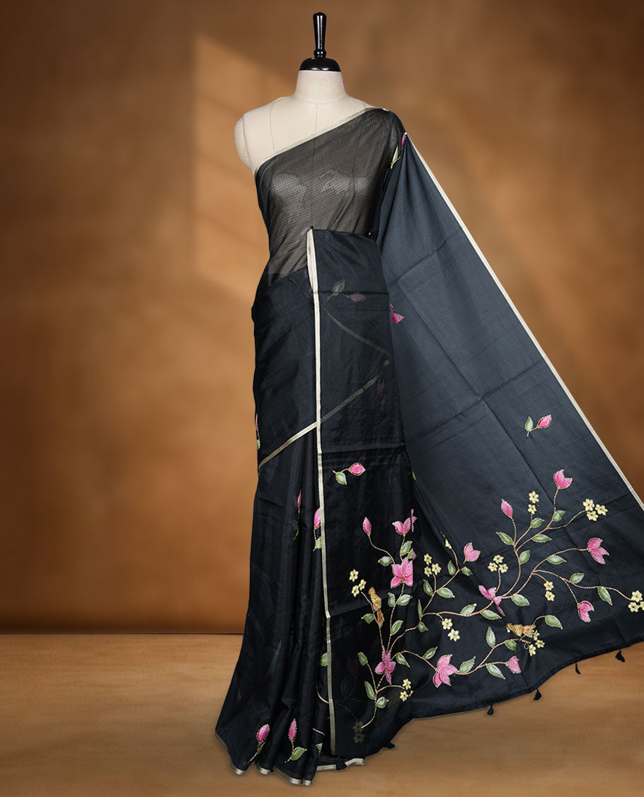 Black colour semi tusser silk saree adorned with fine floral embroidery spread across the body, highlighted by subtle contrast accents along the border, paired with a matching blouse.