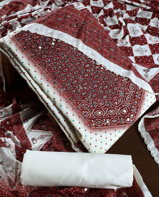 Off white colour churidhar material adorned with mirror work and maroon colour traditional bandhani patterns, paired with a green and maroon colour dotted off white top, plain bottom, and an elegant matching dupatta.