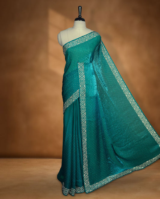 Teal colour chiffon designer saree, featuring delicate scattered stone detailing all over the body, highlighted with intricate stone work and a heavy geometric stone pattern along the border and pallu, paired with a matching embellished blouse.