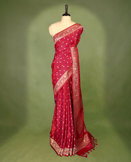 Pinkish red colour semi Banaras satin crepe saree featuring gold zari woven flower motifs across the body, complemented by an intricately designed traditional banarasi border and richly woven pallu, paired with a matching blouse piece.