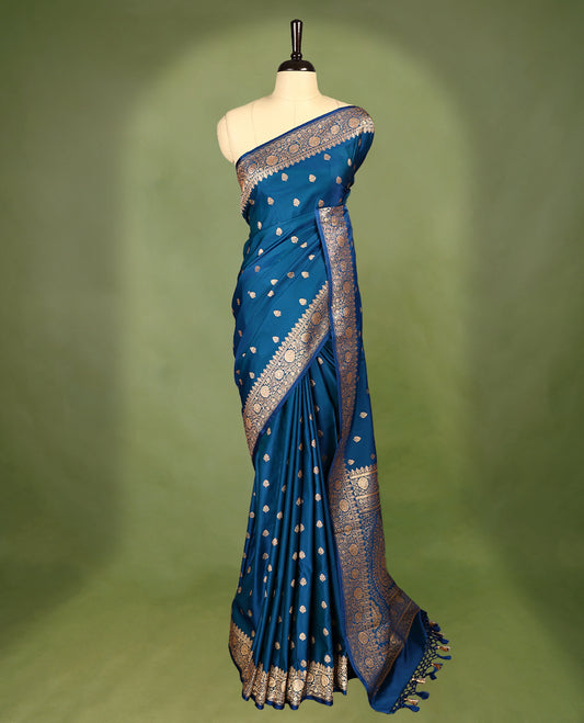 Peacock blue colour semi Banaras satin crepe saree featuring gold zari woven floral motifs across the body, complemented by an intricately designed traditional banarasi border and richly woven pallu, paired with a matching blouse piece.