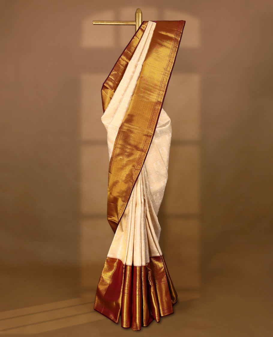 Off-white Colour traditional Kancheepuram silk saree adorned with antique zari floral motifs arranged in a symmetric grid on the body, featuring a contrasting border with floral, peacock, and chakram motifs, and paired with a matching blouse.