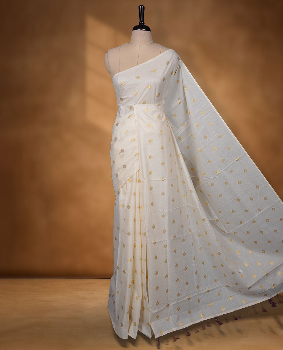 Off-white colour cotton Kerala saree featuring delicate gold butti motifs across the body and pallu, finished with a subtle gold border and tasselled pallu, paired with a matching blouse.