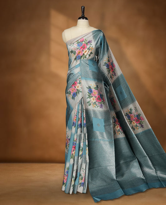 Blue color semi tussar Banarasi saree featuring vibrant multi-color floral prints across the body, with geometric zari patterns, and a matching blouse detailed with silver woven buttas.