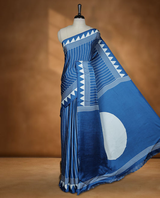 Blue colour semi modal saree highlighted with white triangle border accents and sleek striped detailing across the drape, featuring a bold oversized white circular motif on the pallu, paired with a matching printed blouse.