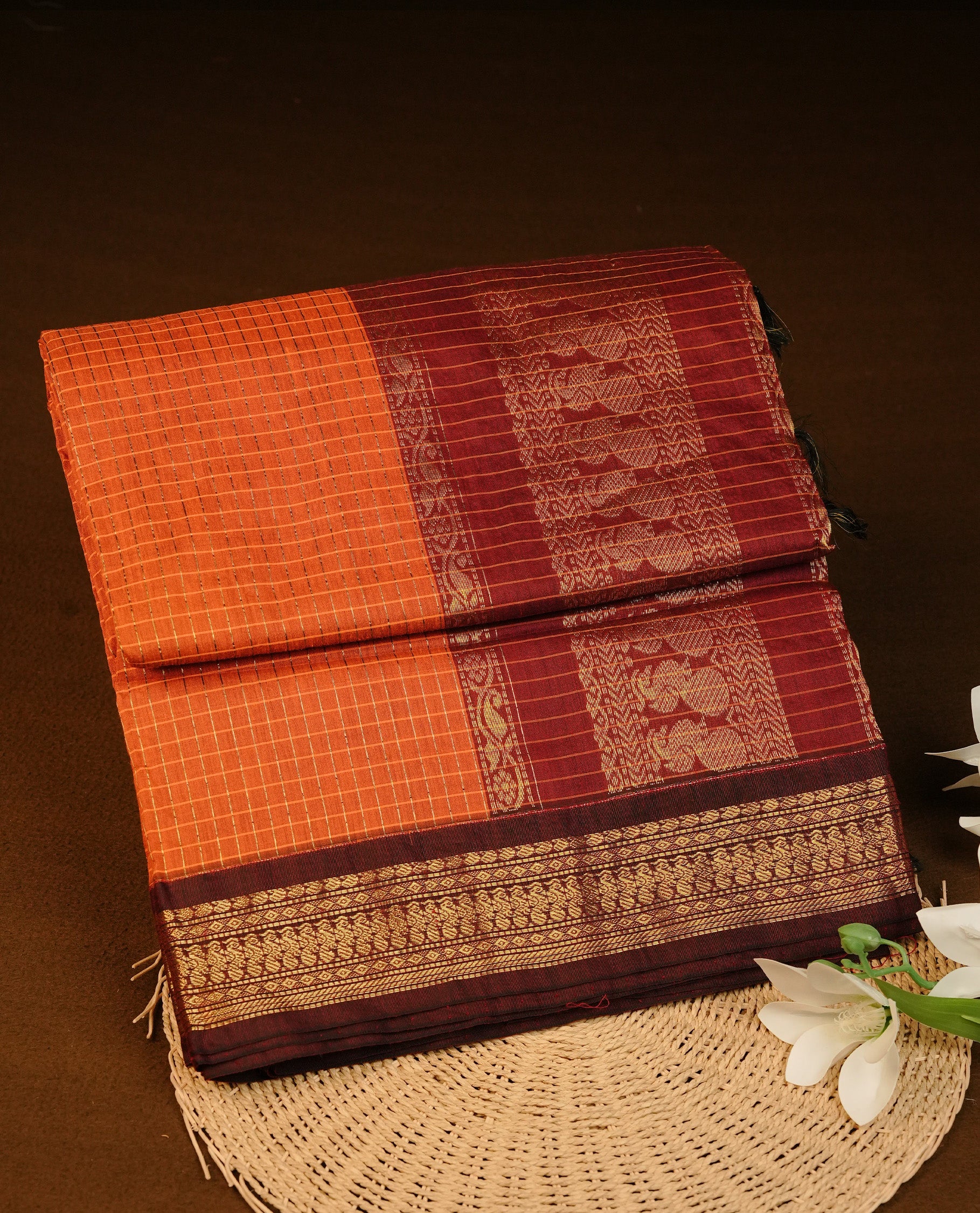 Orange colour Kalyani cotton saree featuring Gold zari check pattern on body, complemented by Contrast Dark Brown colour with grand gold zari border & pallu, matching blouse piece