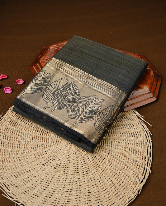 Blackish Grey colour semi tussar silk saree featuring an elegant plain body with traditional gold zari detailed border for a refined look. Comes paired with a matching blouse piece.
