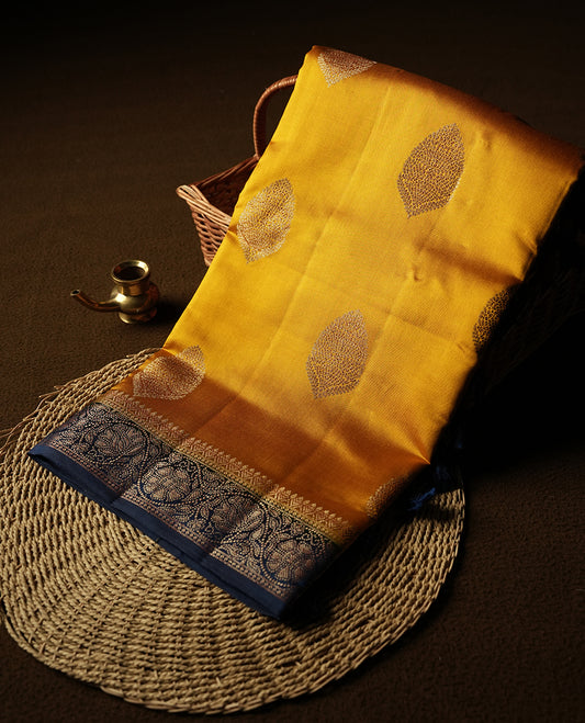 Golden Yellow Colour Banaras Soft Silk Saree with Gold Zari Oval Leaf Buttas, Intricate Multi-Layered Floral Border, Grand Pallu with Ornate Brocade Motifs in Navy Blue and Gold Tones, and Matching Blouse.