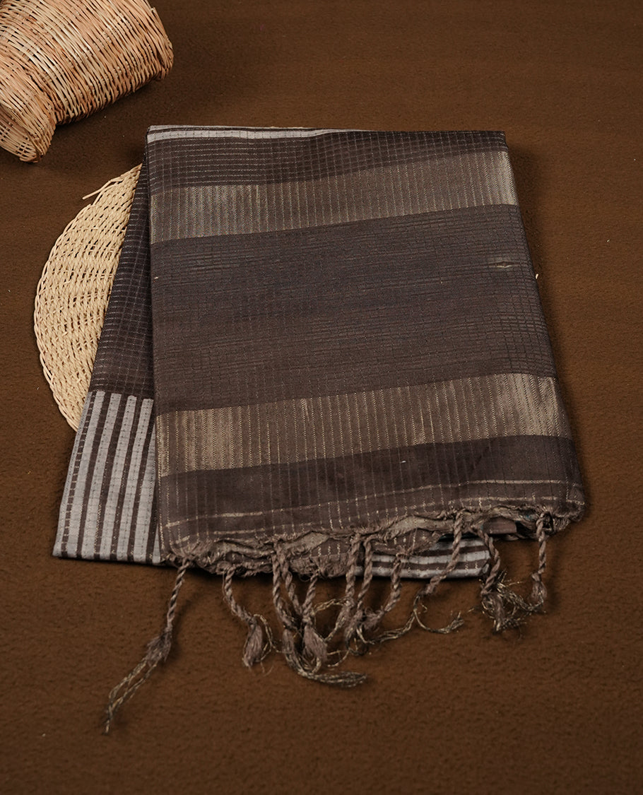 Charcoal Brown colour semi tussar silk saree crafted in breathable cotton, featuring subtle striped textures across the body, a minimalist pallu with fine linear detailing, and a coordinated plain blouse piece.