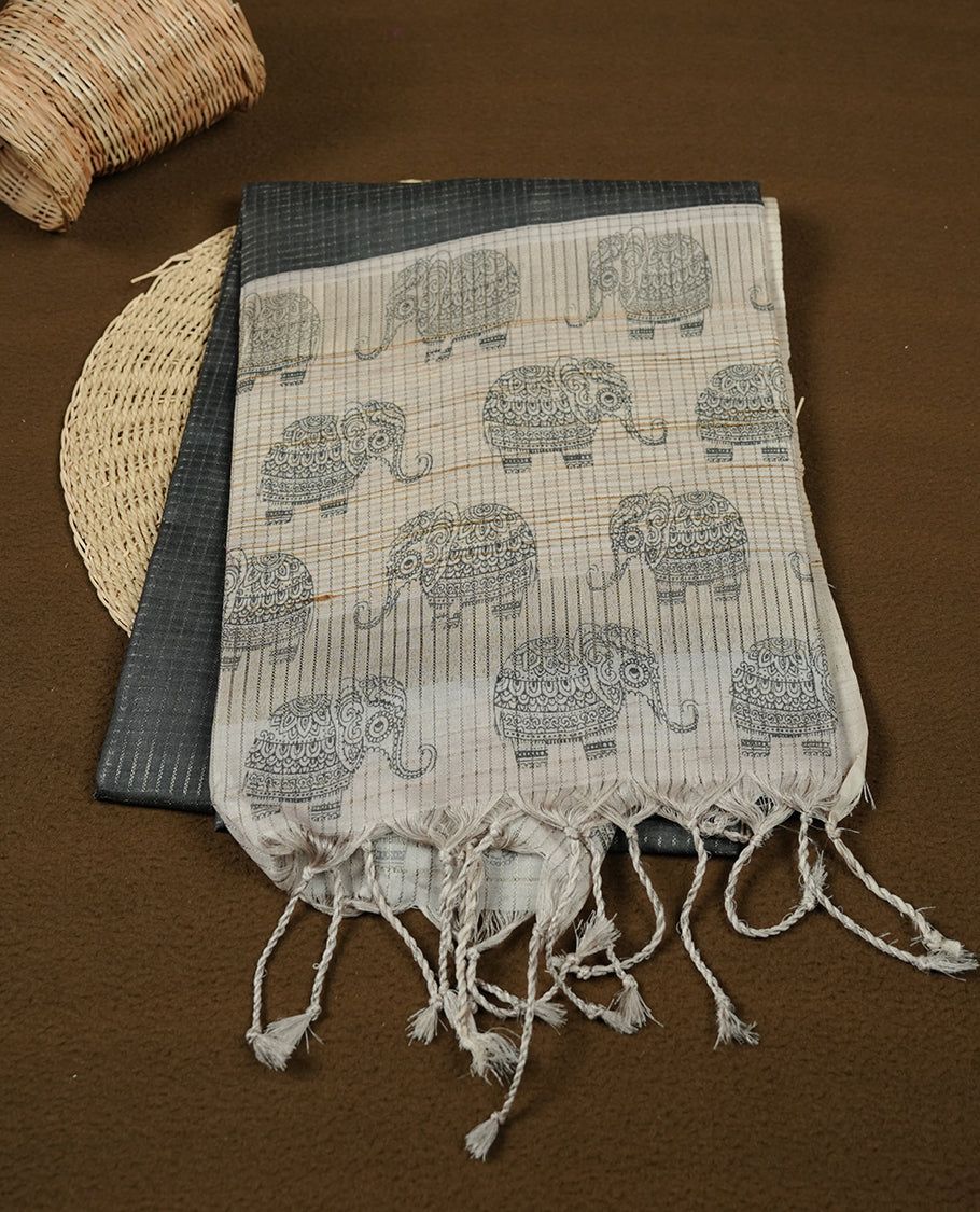 Charcoal green colour semi tussar silk saree featuring an all-over elephant motif print across the body, complemented by a contrasting blue pallu and border, and paired with a matching blouse piece.