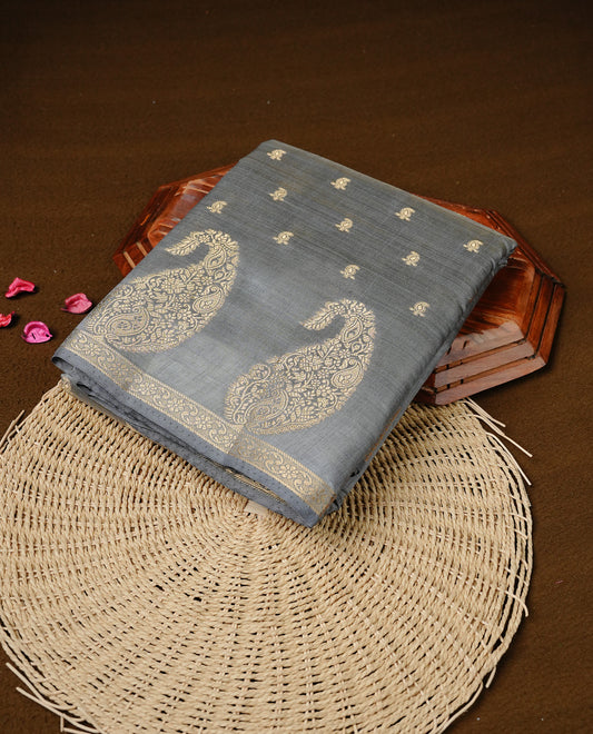 Grey colour semi tussar silk saree featuring small gold zari mango motifs across the body, enhanced with a self colour border showcasing elegant gold zari mango patterns. Paired with a matching blouse piece.