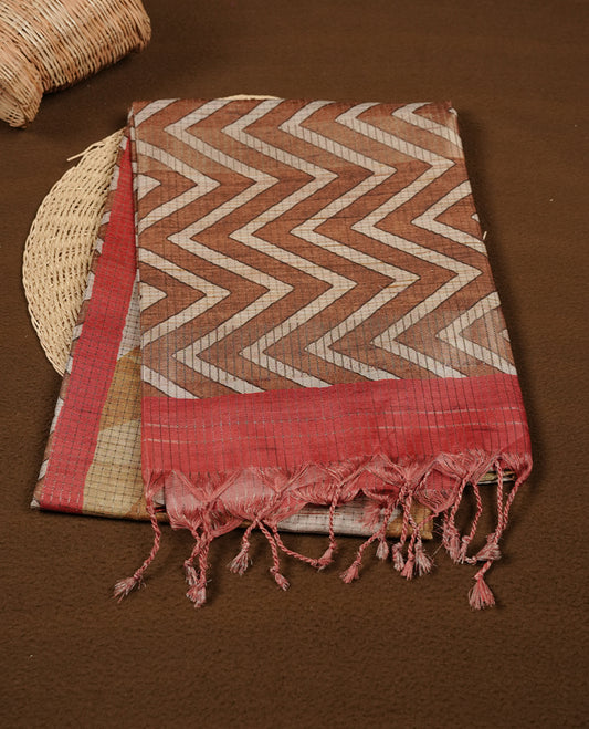 Rust brown colour semi tussar silk saree featuring bold zigzag geometric prints across the body, highlighted with a contrasting coral-red border and detailed pallu, paired with a matching printed blouse piece.