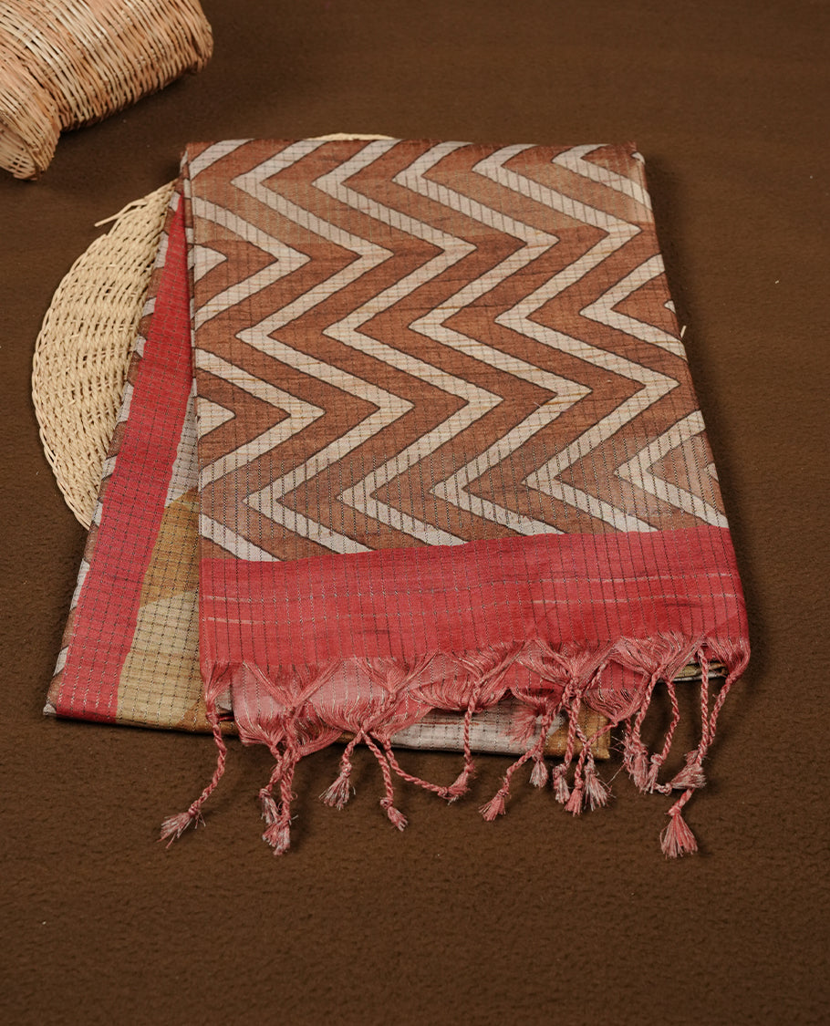 Rust brown colour semi tussar silk saree featuring bold zigzag geometric prints across the body, highlighted with a contrasting coral-red border and detailed pallu, paired with a matching printed blouse piece.