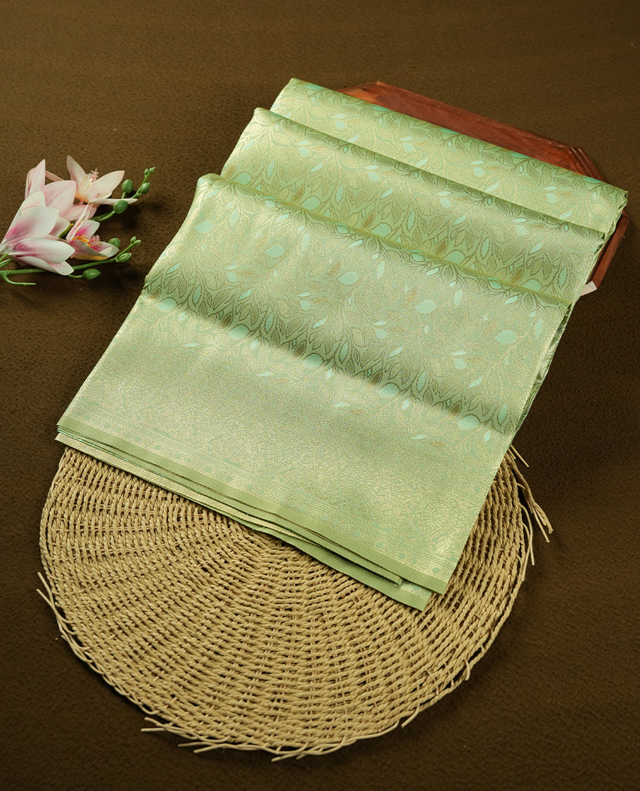 Light Green colour semi soft silk saree featuring an all-over woven leaf pattern in gold colour, finished with a matching border adorned with detailed paisley and floral motifs, and a matching blouse piece.