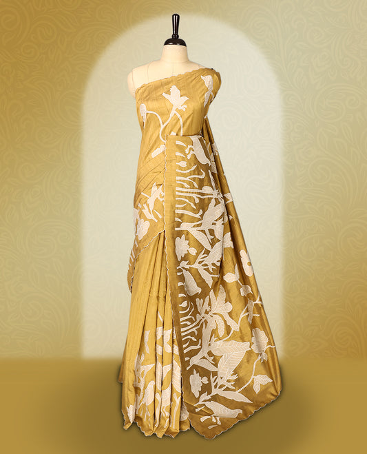 Golden Brown colour semi tussar saree adorned with elegant white applique work in leaf and floral designs across the body and border, paired with a matching blouse.