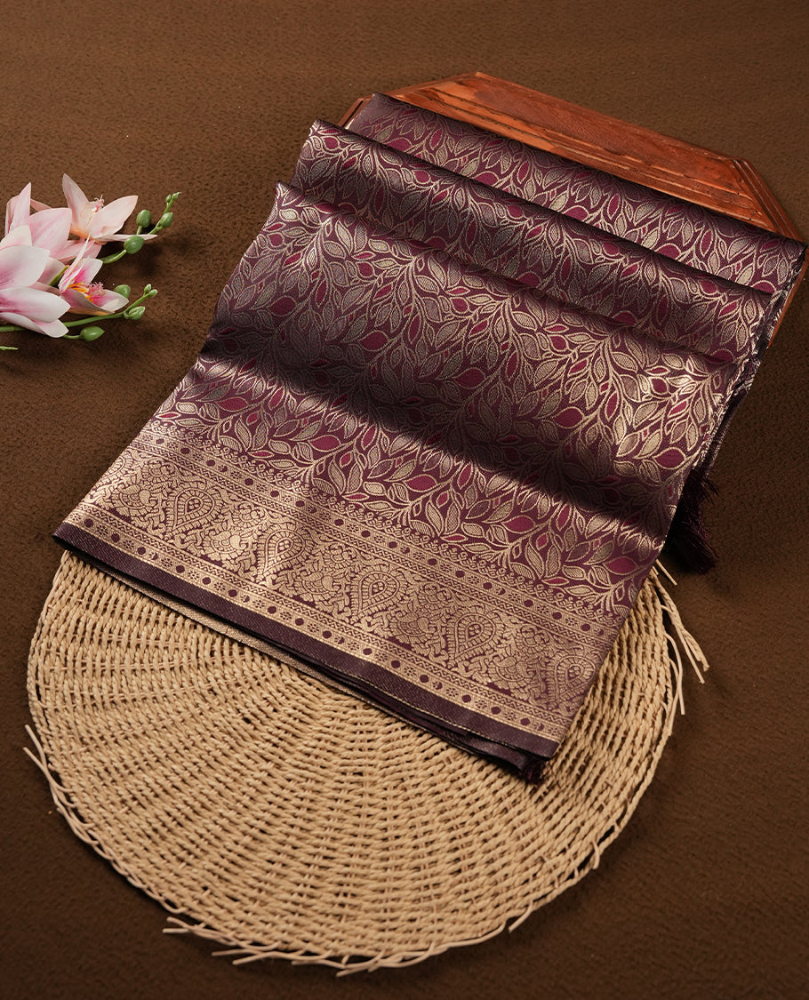 Dark Brown colour semi soft silk saree featuring an all-over woven leaf pattern in gold colour, finished with a matching border adorned with detailed paisley and floral motifs, and a matching blouse piece.