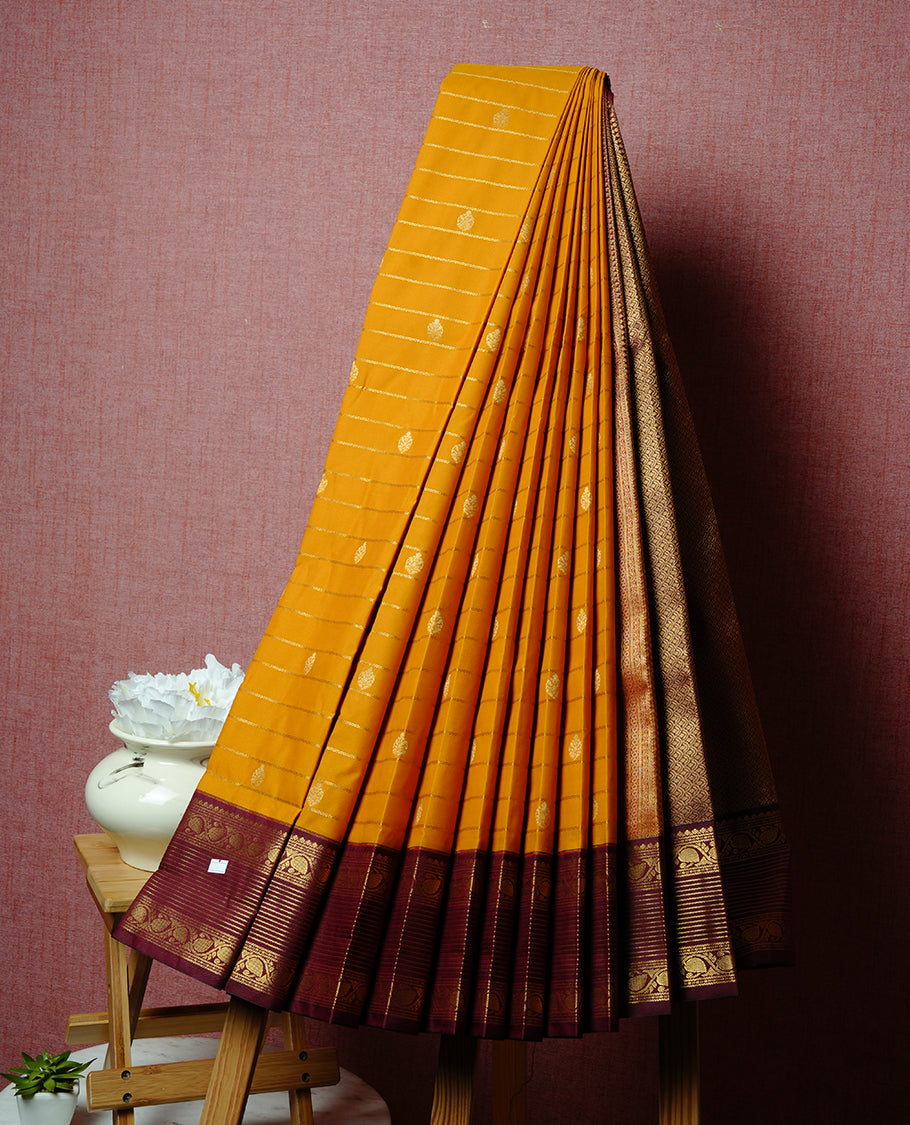 Mustard coloured soft silk saree featuring gold zari stripes and motifs across the body, enhanced with a contrast Maroon border and pallu, paired with a matching contrast blouse piece.