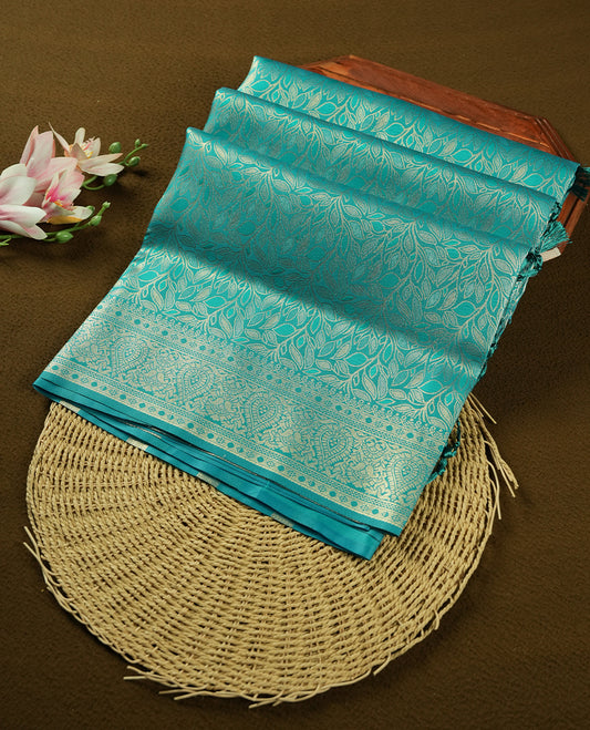 Teal colour semi soft silk saree featuring an all-over woven leaf pattern, finished with a matching border adorned with detailed paisley and floral motifs, and a matching blouse piece.