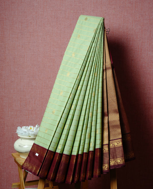 Mint green coloured soft silk saree featuring gold zari stripes and motifs across the body, enhanced with a contrast Maroon border and pallu, paired with a matching contrast blouse piece.